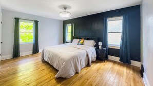 A bedroom with a large bed, two windows with curtains, a bedside table with a lamp, and a ceiling light.