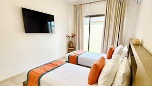 Habitación de hotel con dos camas, un televisor de pantalla plana montado en la pared, una ventana con cortinas y una mesita con un florero.