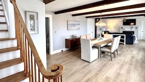 A spacious living area with a wooden staircase, a dining table, and a kitchen in the background.
