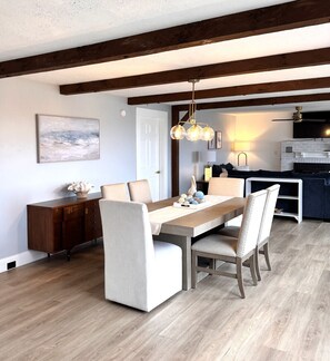 A dining area with a wooden table, chairs, a chandelier, and a kitchen in the background.