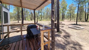 Una terraza de madera con mesa y sillas, un espacio cubierto y un pozo de fuego en un camping rodeado de árboles.