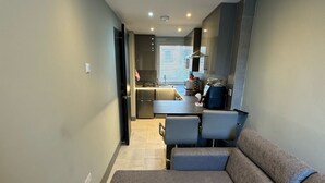 En moderne stue med en hjørnesofa, et køkkenområde med et vindue og indbyggede loftslamper.