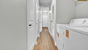 A laundry room with white appliances, a wooden floor, and a hallway leading to a door.
