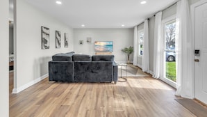 A modern living room with a dark grey sectional sofa, wooden flooring, white walls, and a large window with curtains.