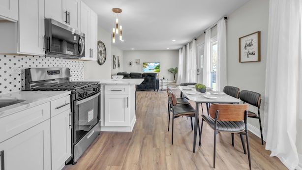 A modern kitchen with white cabinetry, a built-in microwave, and a stove. There is a dining area with a table and chairs, and a large window providing natural light.