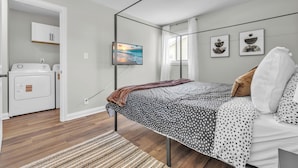 A bedroom with a bed, a television, a window with curtains, and a washing machine in the adjacent room.
