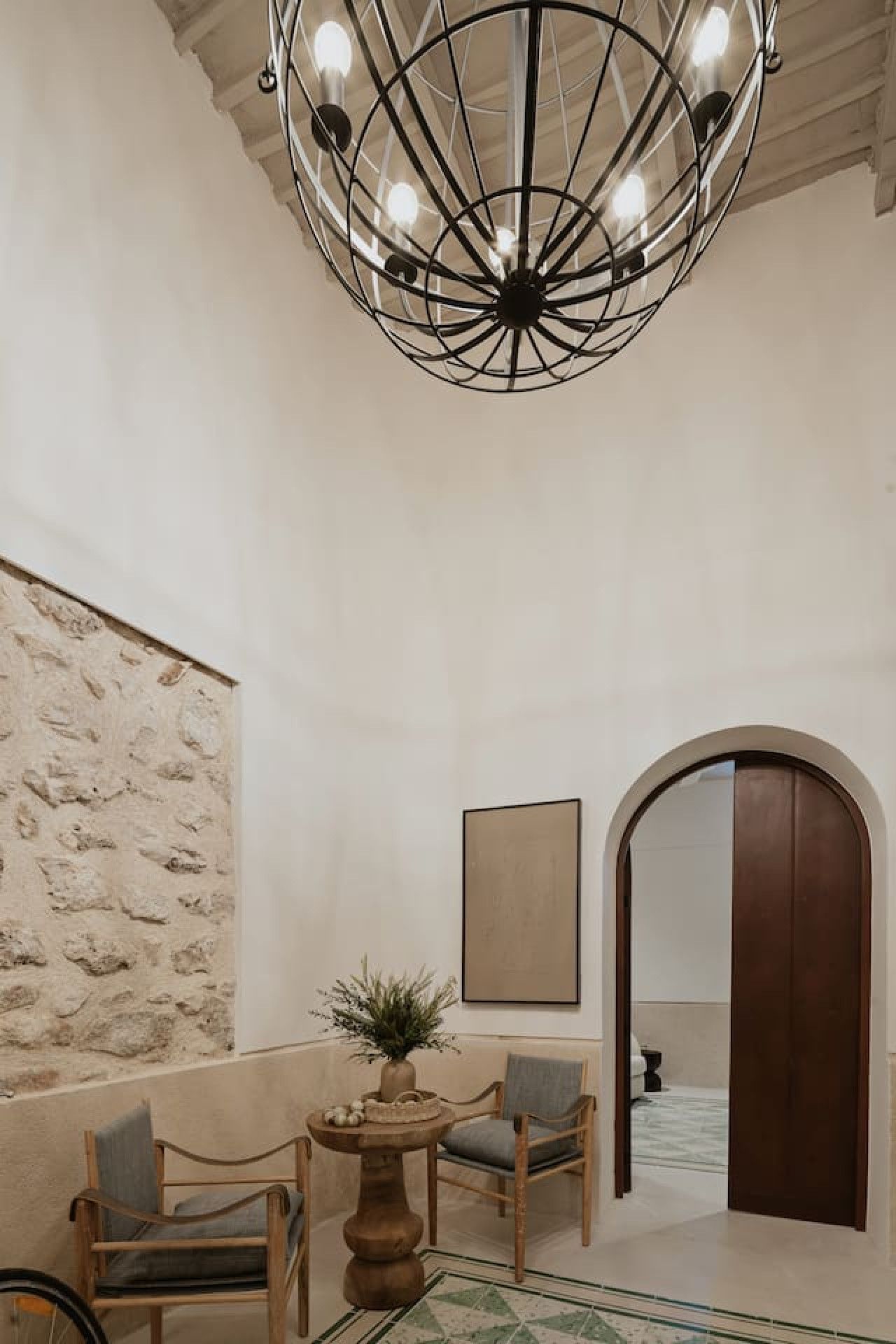 Un vestíbulo con pared de piedra, una mesa redonda con una planta, dos sillas, una gran lámpara de araña y una puerta de madera que da a otra habitación.