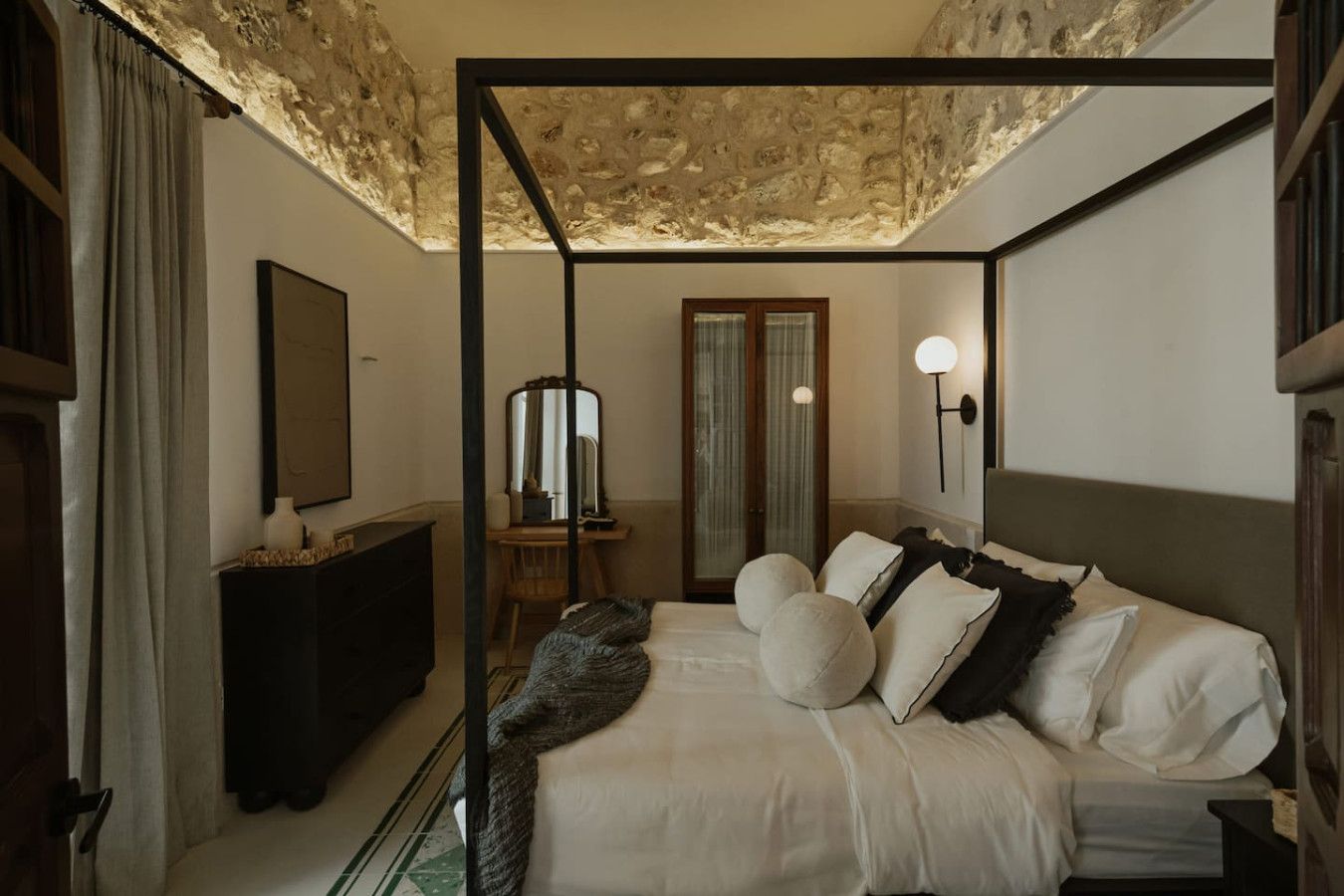 A four-poster bed with a stone ceiling, a mirror, a bedside table, and a lamp.