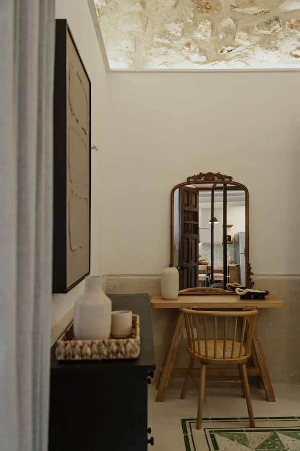 A room with a wooden desk, a mirror, a framed picture on the wall, and a small cabinet with a cushion.