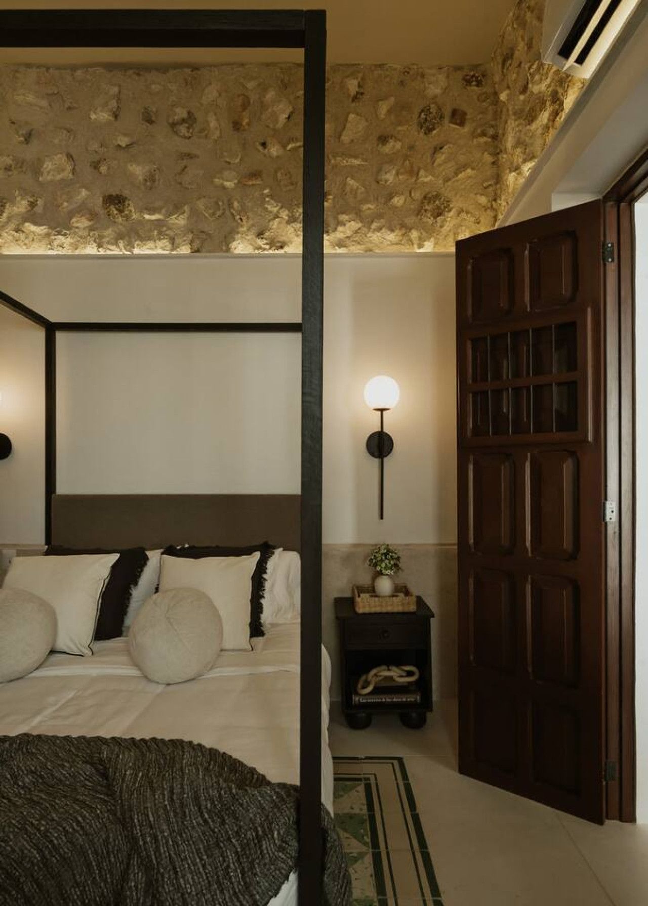 Una camera da letto con soffitto in pietra, un letto con biancheria da letto bianco e scuro, un comodino con una lampada e una porta in legno.