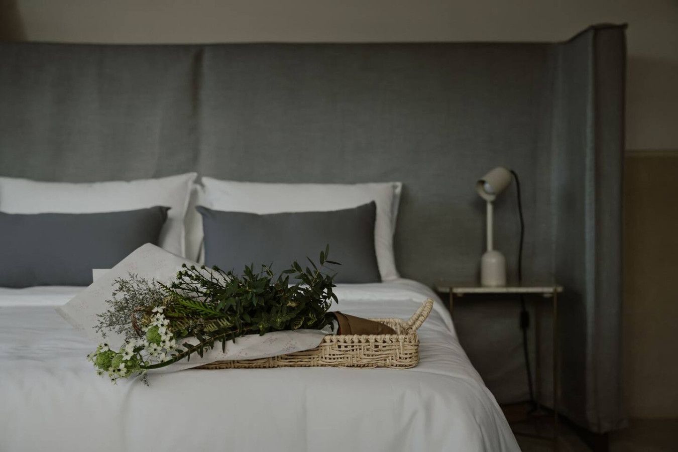 A neatly made bed with white bedding, a woven basket with greenery, and a bedside lamp on a nightstand.