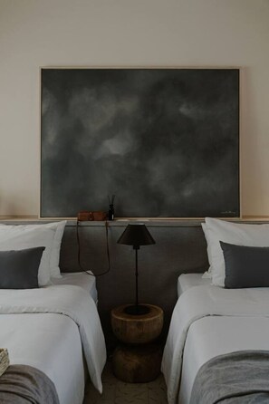 Une chambre d’hôtel avec deux lits, un grand tableau abstrait accroché au mur, un petit tabouret en bois et une lampe de sol noire.