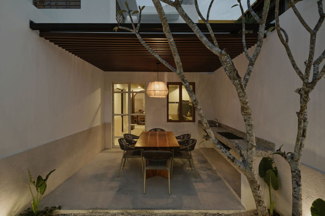 A modern outdoor dining area with a wooden table and chairs, surrounded by a low wall and a tree.
