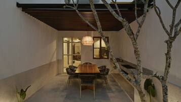 A modern outdoor dining area with a wooden table and chairs, surrounded by a low wall and a tree.