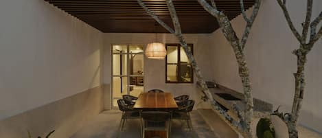 A modern outdoor dining area with a wooden table and chairs, surrounded by a low wall and a tree.