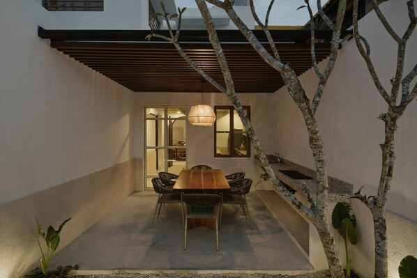 Un espacio exterior moderno para comer, con una mesa y sillas de madera, rodeado por un muro bajo y un árbol.