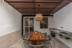 A covered patio with a wooden table and wicker chairs, a hanging light fixture, and a kitchen area with a sink and countertop. - Casa Meraki Mérida - Paseo de Montejo (Centro)