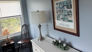 A room with a framed collage on the wall, a lamp, a chair, a small table with a vase, and a window with blinds. - Charming 2-bedroom condo with WiFi, AC in gorgeous Kitty Hawk (Kitty Hawk)