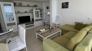 Un salon avec un canapé vert, un fauteuil blanc, une table basse, un téléviseur et un meuble encastré.