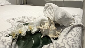 Un lit bien fait, avec une couverture à motifs, une serviette enroulée, des fleurs blanches et un coquillage.