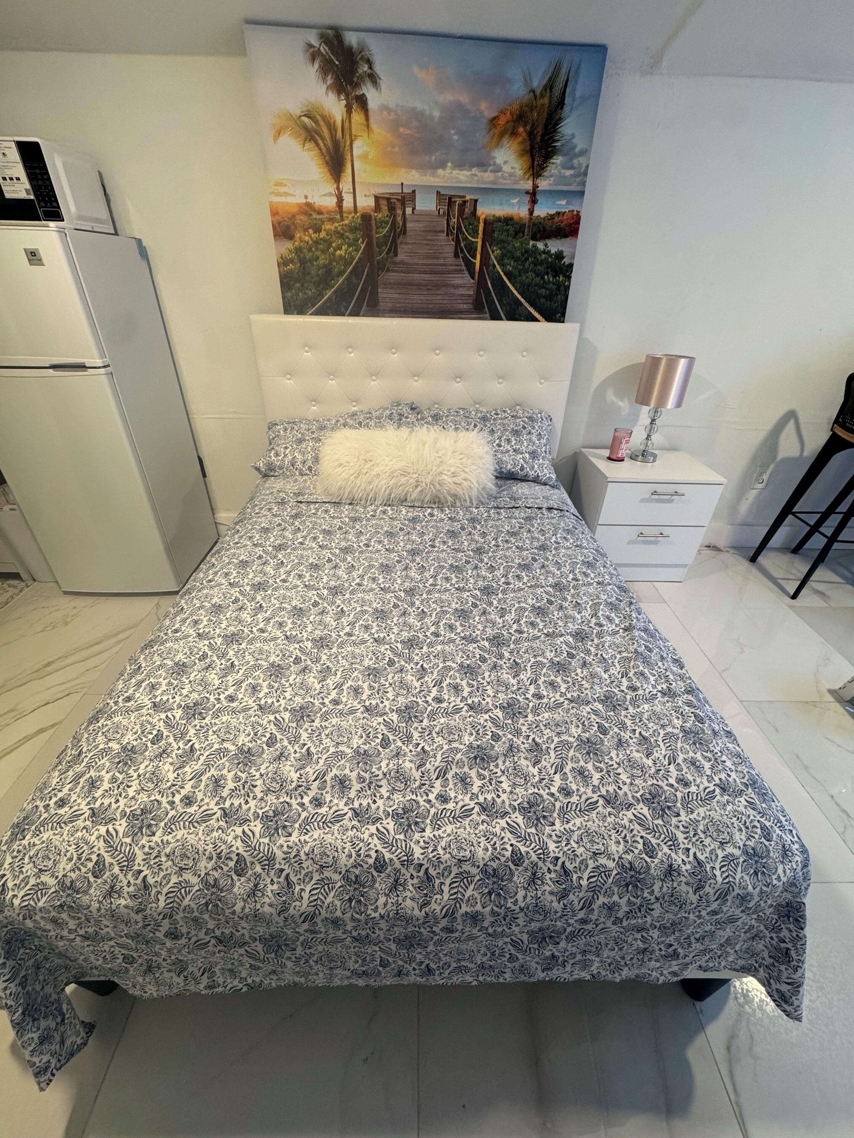 Quarto com cama, mesa de cabeceira, luminária, geladeira e uma pintura de uma cena de praia.