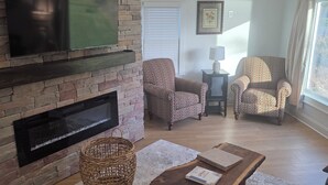 Ein Wohnzimmer mit Kamin aus Stein, zwei gemusterten Sesseln, einem Holztisch und einem flachbildfernseher, der an der Wand befestigt ist.