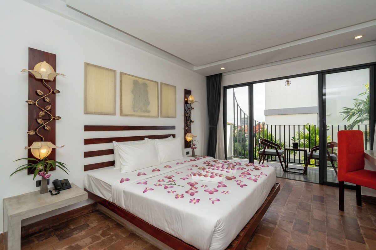 Habitación de hotel con una cama grande, un balcón con sillas y vista a zonas verdes.