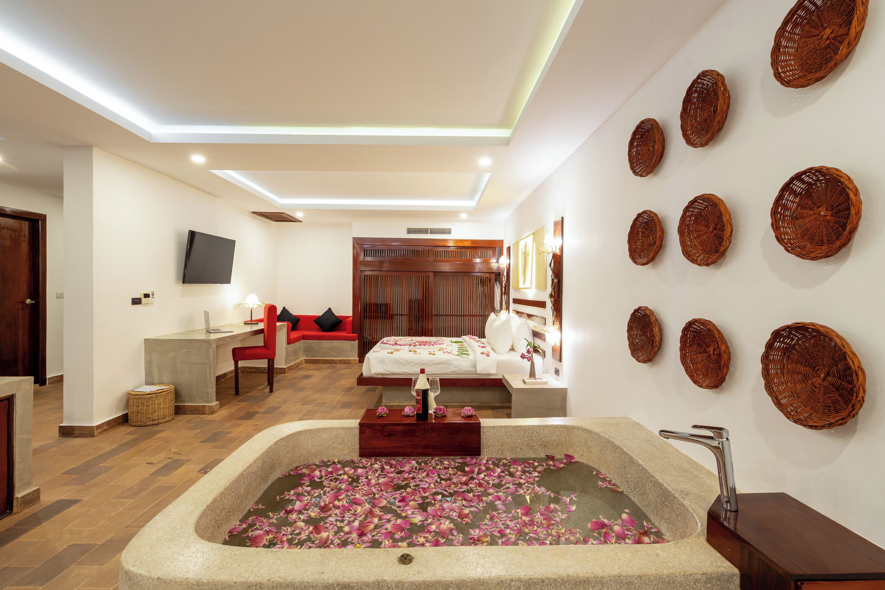 Una habitación de hotel moderna con una bañera grande llena de pétalos de rosa, un cabecero de madera, un televisor de pantalla plana, un escritorio con una silla y cestas decorativas tejidas en la pared.