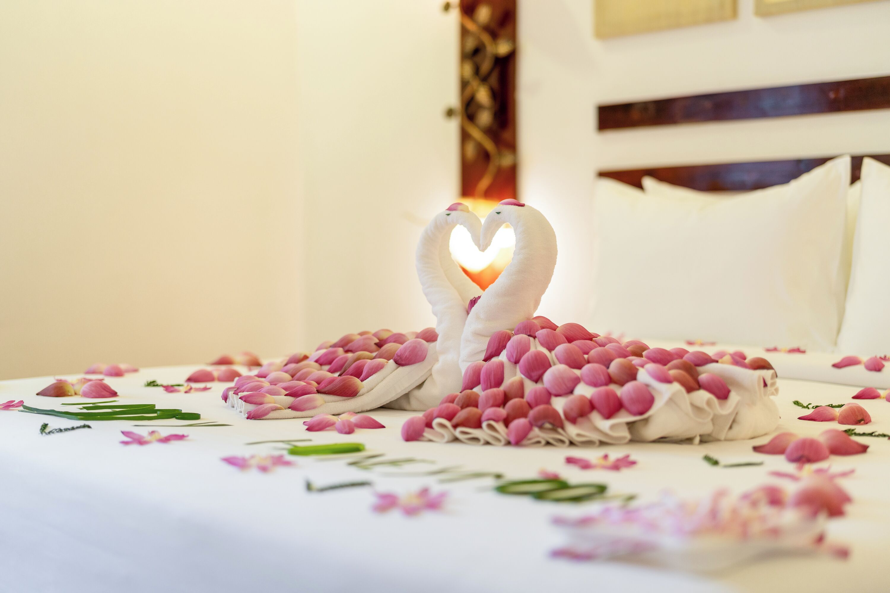 A bed with a heart-shaped towel arrangement and scattered rose petals.
