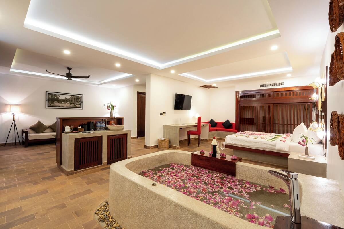 Une chambre d’hôtel moderne avec une grande baignoire remplie de pétales de roses, un bureau avec une chaise, un canapé et un téléviseur fixé au mur.