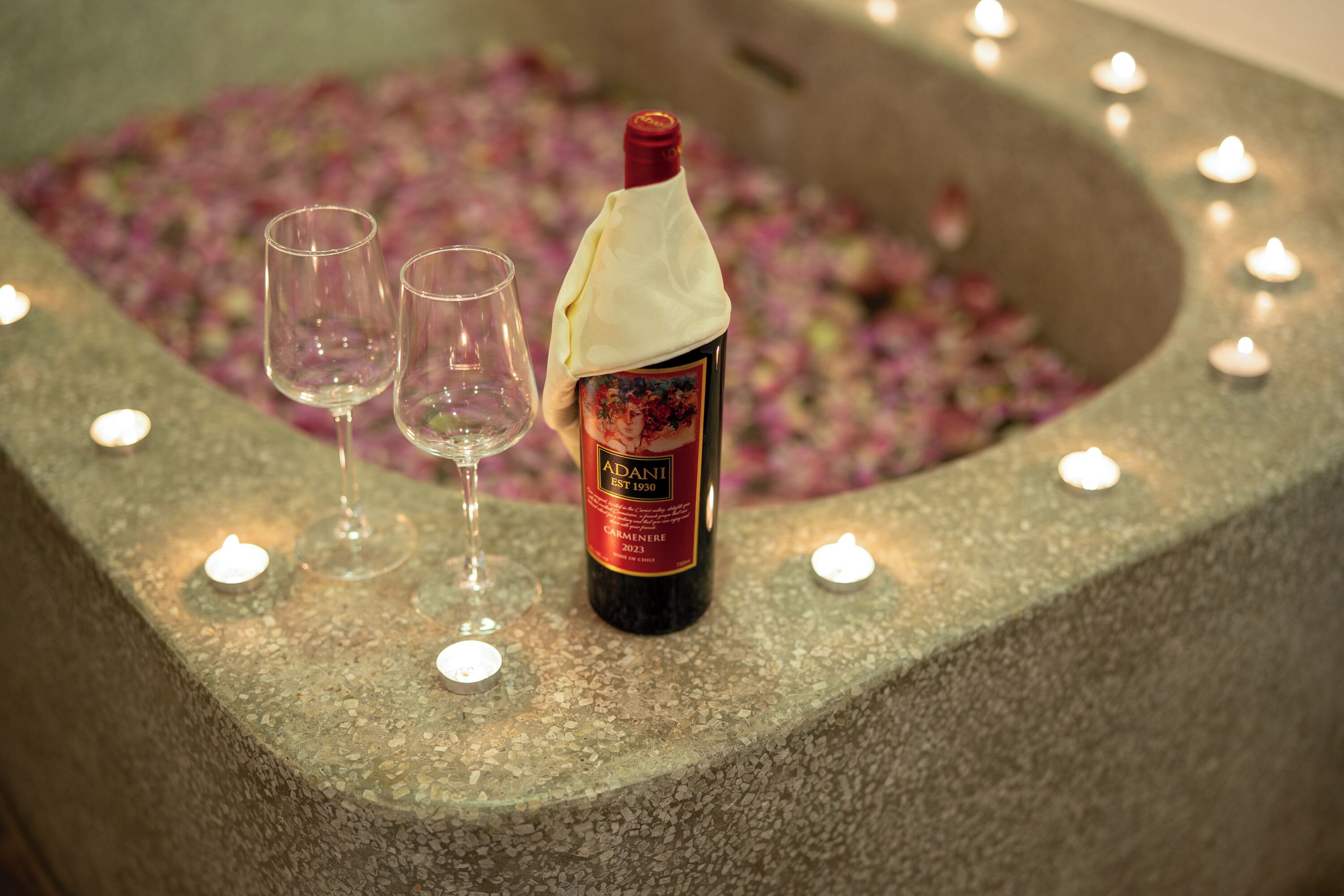 A bottle of Adani wine, two glasses, and candles on a stone surface with flower petals.