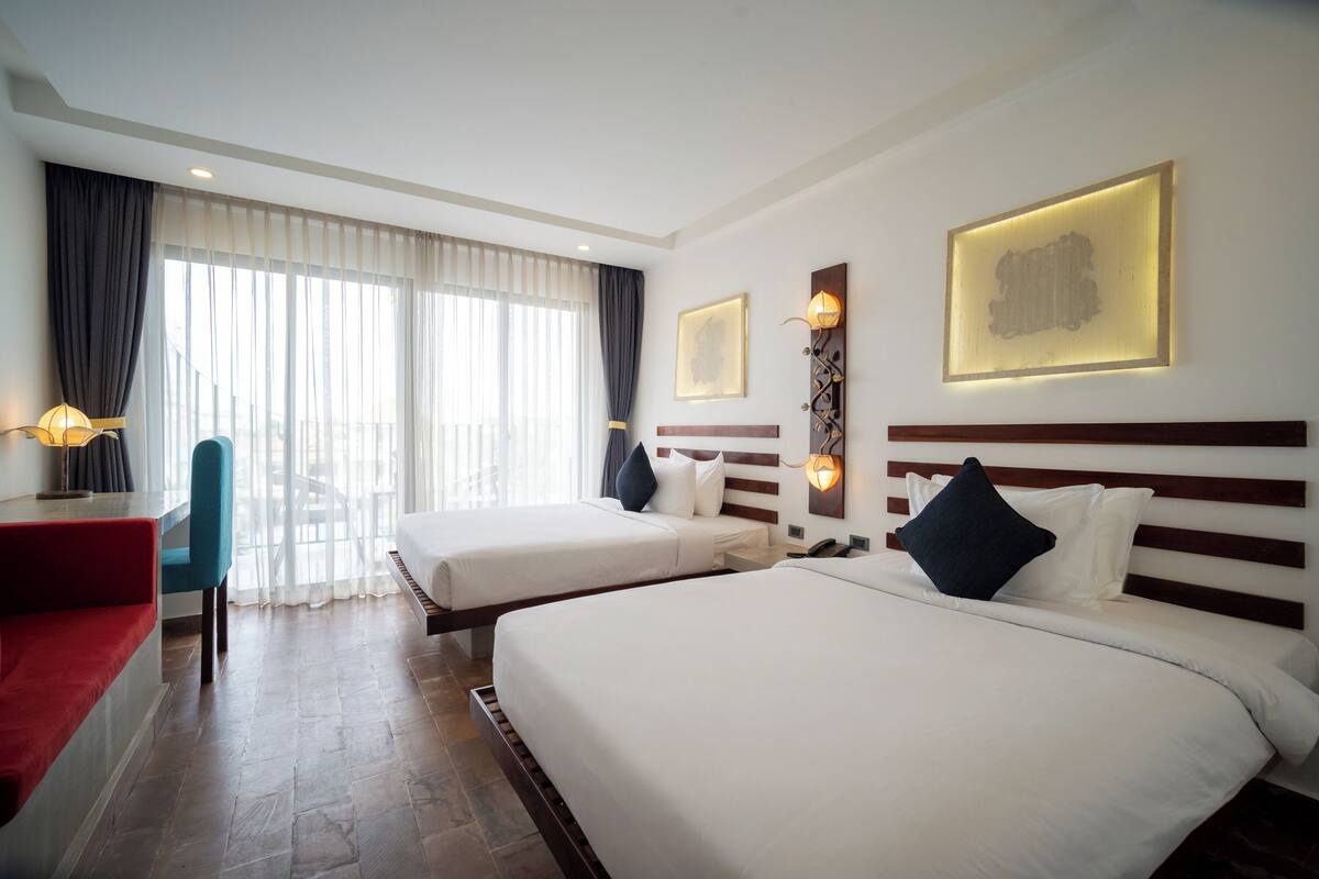 Une chambre d’hôtel avec deux lits, un bureau, une chaise et de grandes fenêtres avec des rideaux.