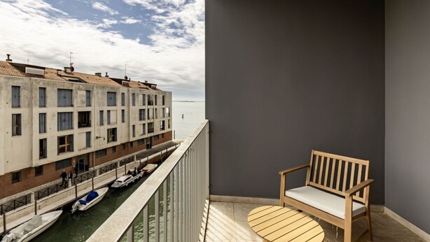 Un balcon avec une chaise en bois, une vue sur un canal avec des bateaux, et un bâtiment situé de l’autre côté de l’eau.