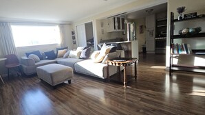 Een ruime woonkamer met een grote bank, voetenbankjes en een salontafel. Er is een eethoek met een tafel en stoelen. De kamer heeft houten vloeren en een keuken op de achtergrond.