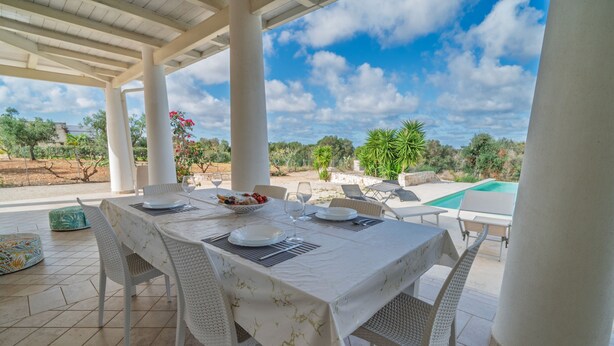 Ein Essbereich im Freien mit Tisch für vier Personen, umgeben von weißen Korbstühlen, unter einer überdachten Terrasse mit Blick auf einen Pool und Grünflächen.