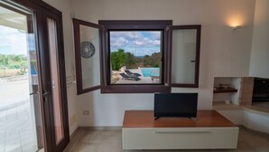 Ein modernes Wohnzimmer mit Fernseher auf einem Holztisch, einem Kamin und Blick auf Pool und Grünfläche.