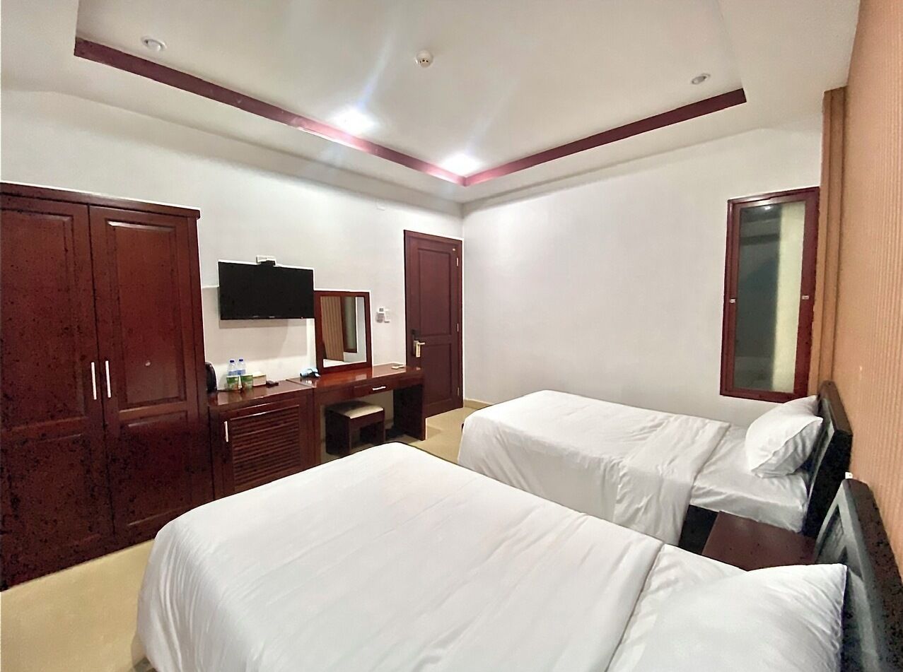 Una habitación de hotel con dos camas individuales, un armario de madera, un escritorio con silla, un televisor de pantalla plana montado en la pared y un espejo sobre la puerta.