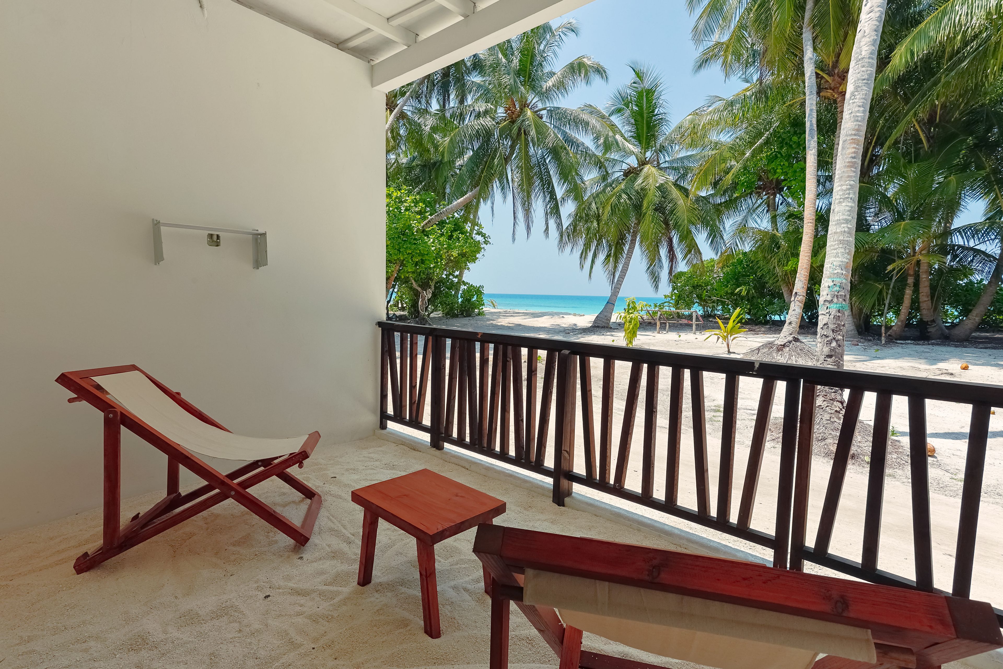 A balcony with a wooden bench, a small table, and a wooden chair. The balcony overlooks a beach with palm trees and the ocean.