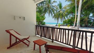 A balcony with a wooden bench, a small table, and a wooden chair. The balcony overlooks a beach with palm trees and the ocean.