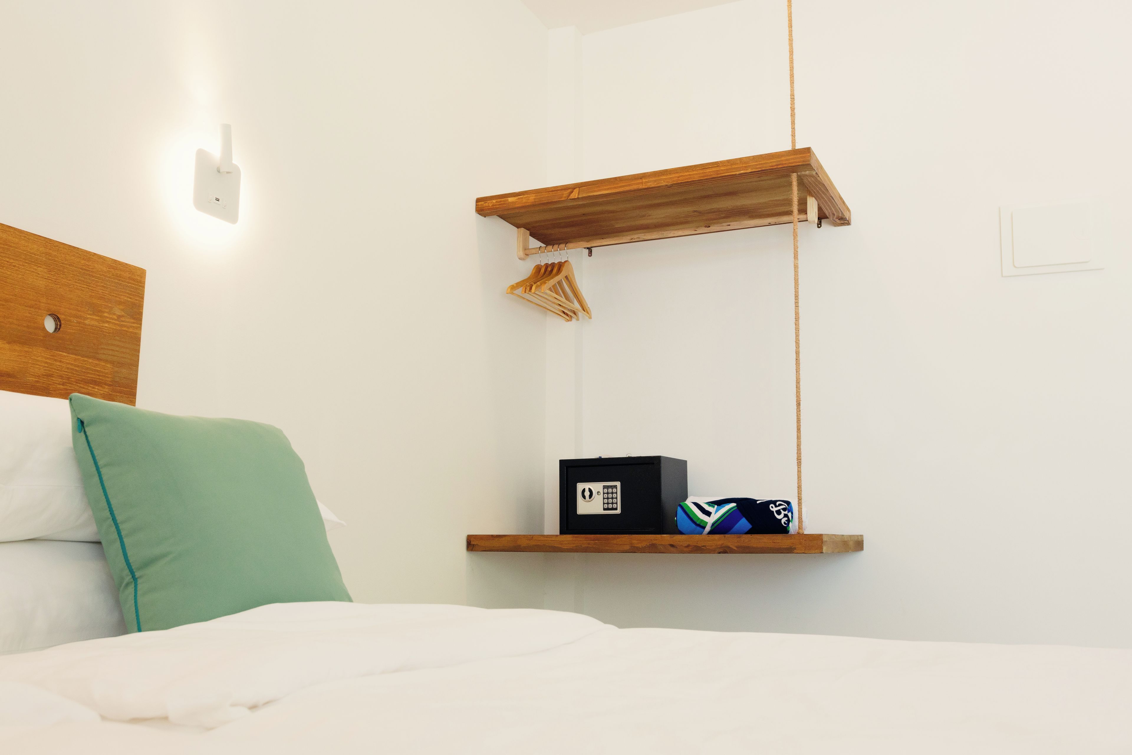 Un dormitorio con una cama, un almohadón verde, un cabecero de madera, un estante fijado a la pared con una caja fuerte negra y una pelota de voleibol, y una lámpara colgante.