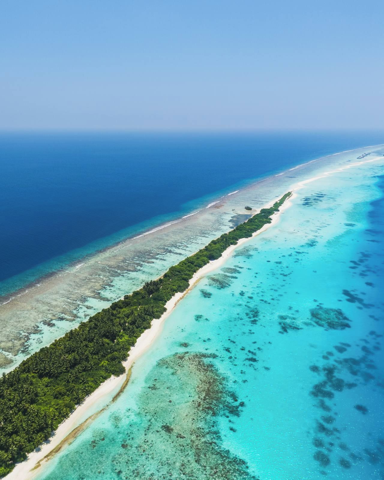 An aerial view of a coastal area with a long stretch of beach, clear turquoise waters, and dense green vegetation along the shoreline.