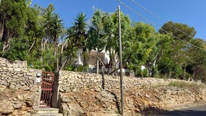 A stone wall with a gate, a paved road, and a building with trees in the background.