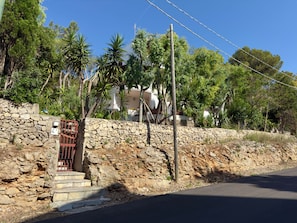 A stone wall with a gate, a paved road, and a building with trees in the background.