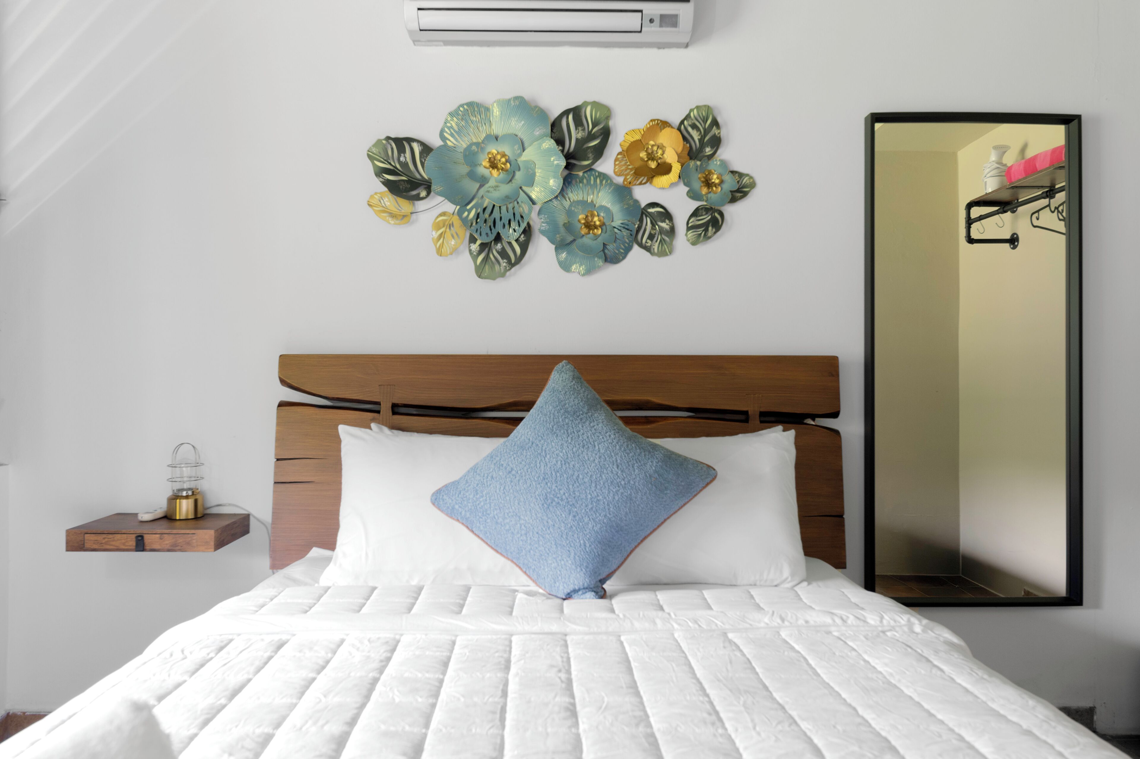 Un dormitorio con cabecera de madera, colcha blanca acolchada, almohada azul, aire acondicionado instalado en la pared y un arreglo floral decorativo.
