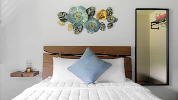 Un dormitorio con cabecera de madera, colcha blanca acolchada, almohada azul, aire acondicionado instalado en la pared y un arreglo floral decorativo.