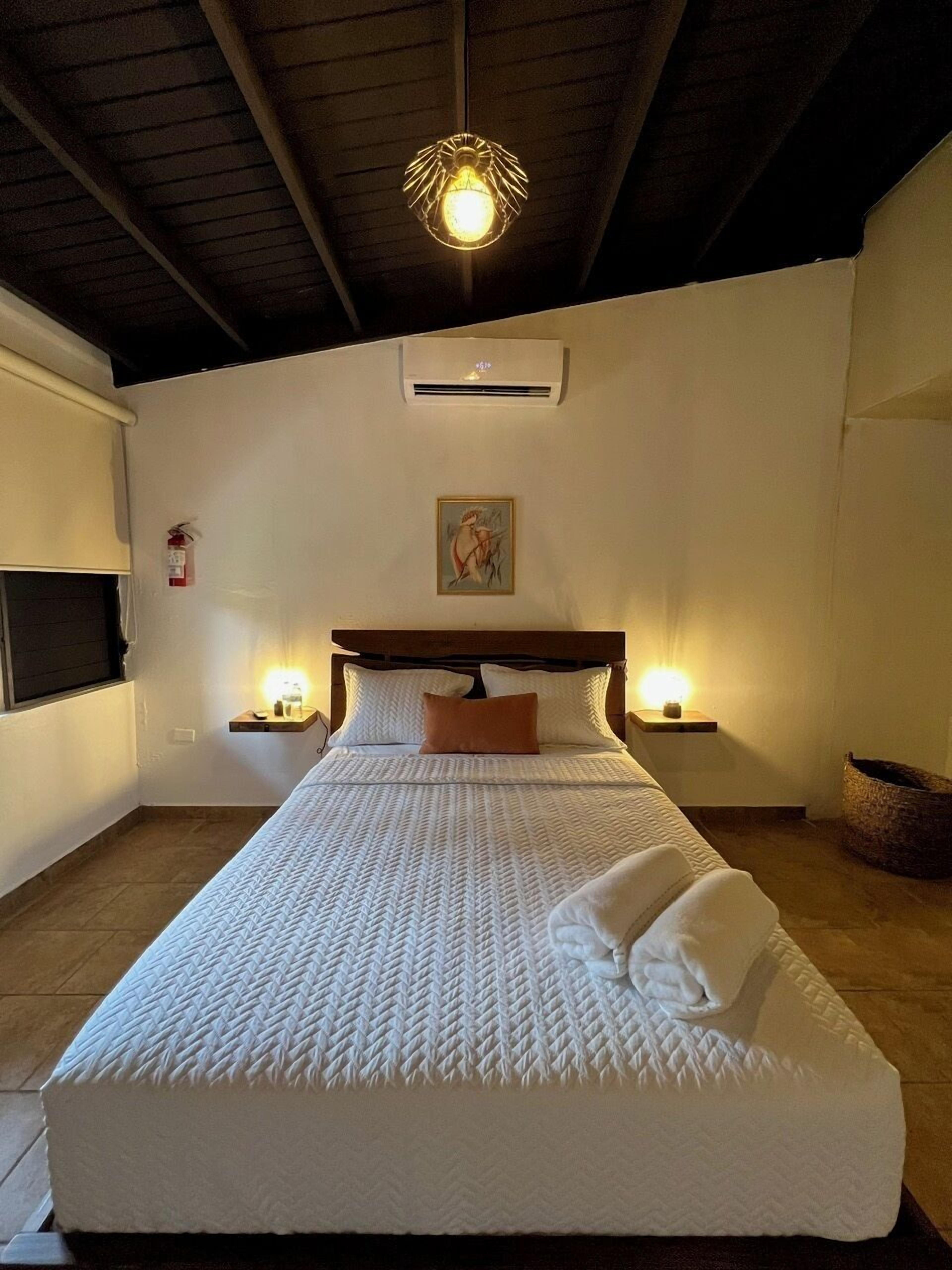 A bedroom with a large bed, two bedside tables with lamps, a ceiling light fixture, an air conditioning unit, and a framed artwork on the wall.