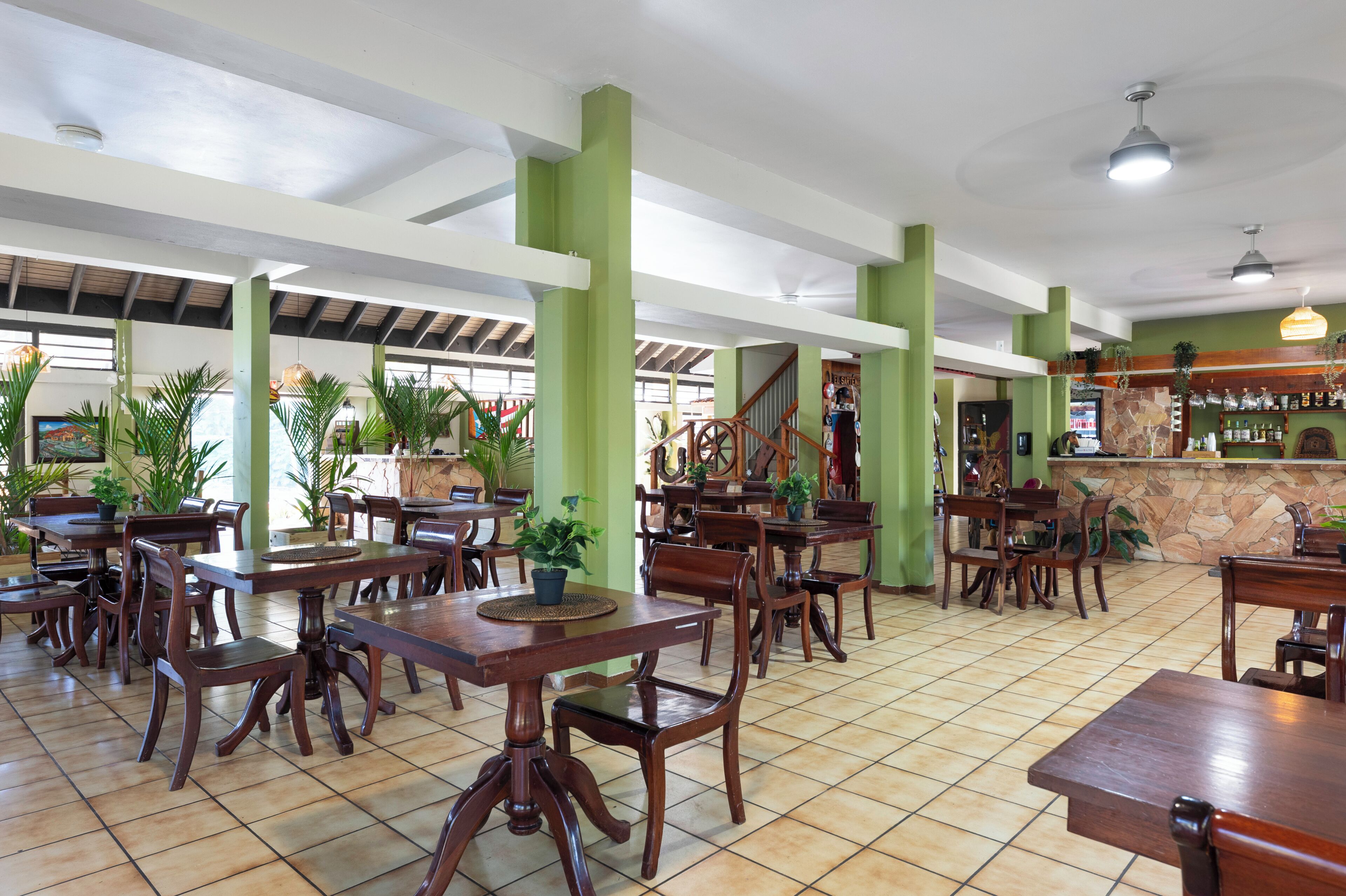 A spacious indoor dining area with wooden tables and chairs, green pillars, and a bar area with a stone counter.