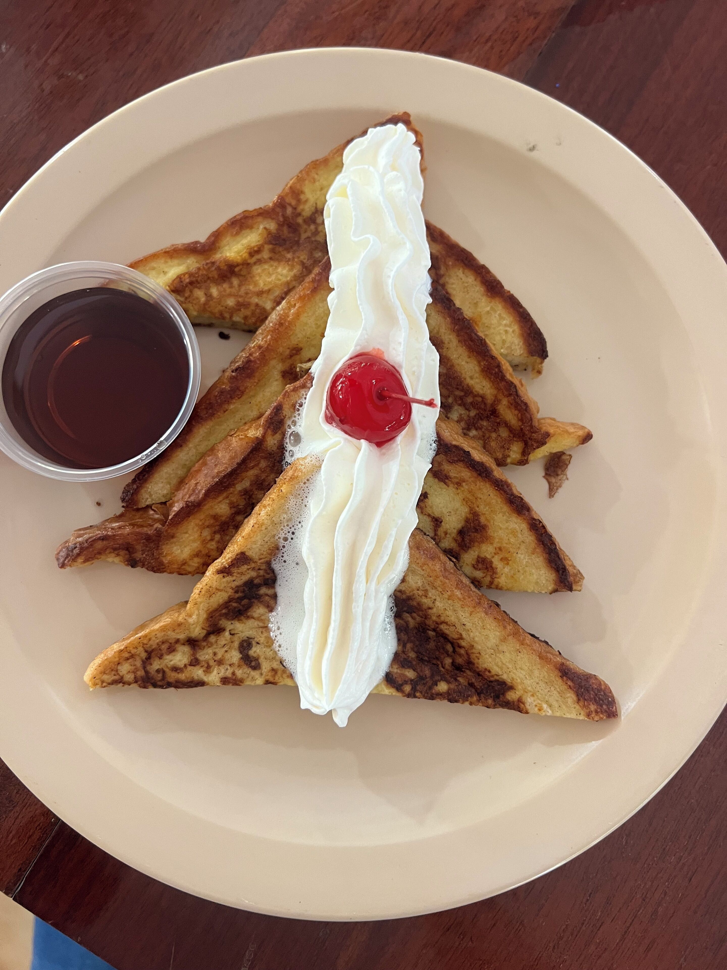 Un plato con tostada francesa, crema batida y una cereza encima, acompañado de una taza pequeña de almíbar.