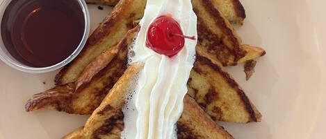 Un plato con tostada francesa, crema batida y una cereza encima, acompañado de una taza pequeña de almíbar.