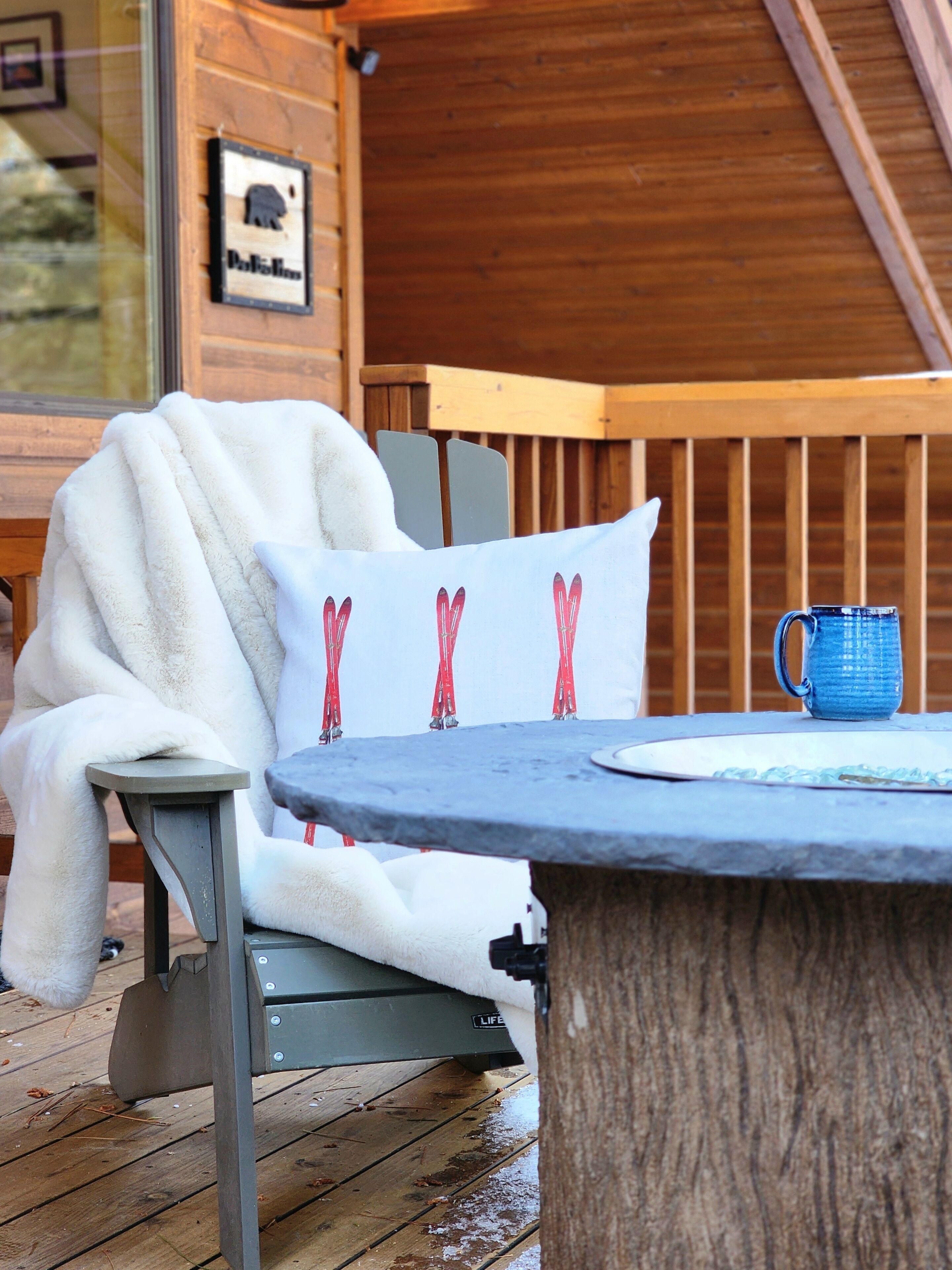 A cozy outdoor seating area with a wooden deck, a white fur-covered chair, a bench, and a table with a blue mug.